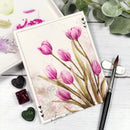 Painting of pink tulips on a white card with art supplies and greenery.
