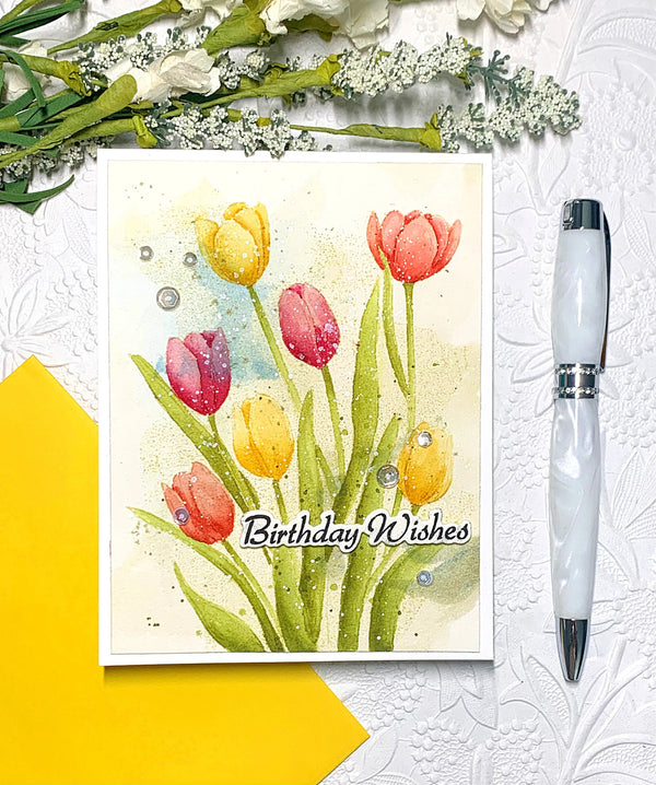 Greeting card with tulips and 'Birthday Wishes' text, pen, and flowers on a textured surface.