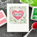 Card with pink heart and 'I'm cheering for you!' text, surrounded by crafting supplies on a dark surface.