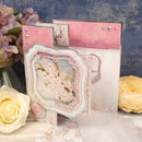 Decorative card with floral design and pink accents, surrounded by flowers on a textured surface.