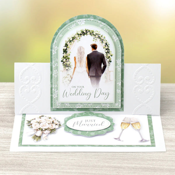 Wedding card with a couple under a floral arch, text 'On Your Wedding Day' and 'Just Married', on a wooden surface.