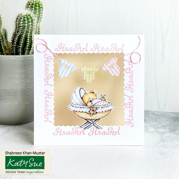 Baby shower card with teddy bear illustration and 'It's a Girl' text, placed on a white surface with a cactus plant in the background.