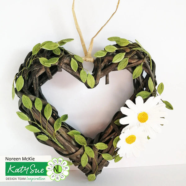Heart-shaped wreath with green leaves and a white flower on a white background, featuring Katy Sue Design Team logo.