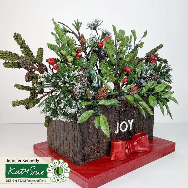 Decorative floral arrangement with greenery, pinecones, and berries on a wooden block with 'JOY' text, on a white background.