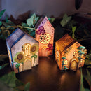 Three decorative paper houses with illuminated interiors, surrounded by green leaves.