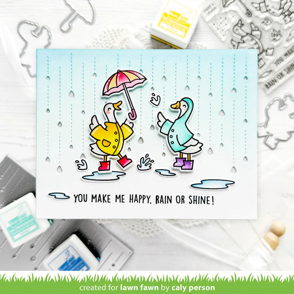 Card with two ducks holding an umbrella and rain boots, surrounded by stamping supplies.