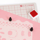 Pink cutting mat with grid and decorative patterns, featuring a red stamp and black stamps on a white background.