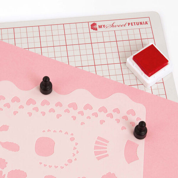 Pink cutting mat with grid and decorative patterns, featuring a red stamp and black stamps on a white background.