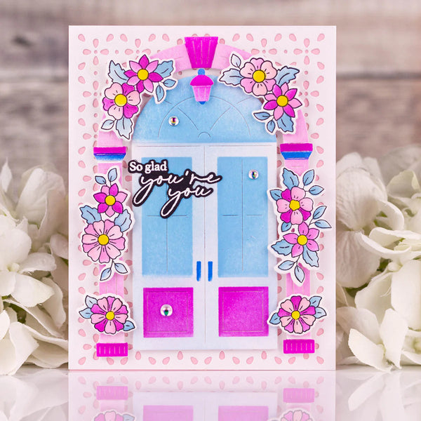 Greeting card with a blue door, pink flowers, and 'So glad you're here' text on a floral background.