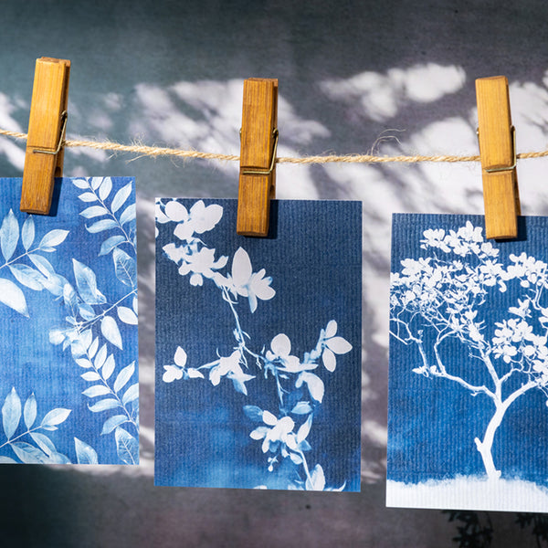 Three blue cards with white floral designs hanging on a string with wooden clips against a gray background.