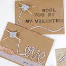 Two brown paper cards with yarn and knitting needles, one saying 'Wool you be my Valentine' and the other 'Love you &', on a white background.
