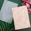Two patterned notebooks, one gray and one pink with floral design, on a green surface with flowers and leaves.