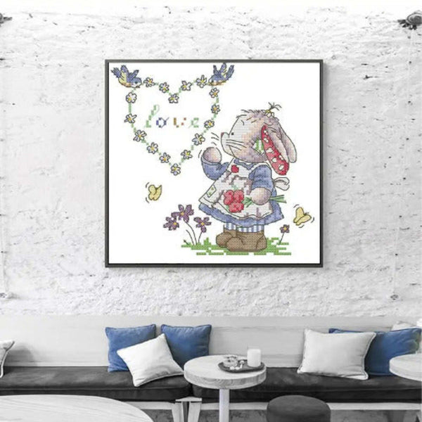 Framed cross-stitch art of a bunny holding flowers with a heart-shaped 'love' sign on a white brick wall.