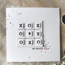 Card with tic-tac-toe design featuring 'XO' and 'O' symbols, with a heart in the center, on a textured surface.