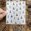 Metal stencil with tree patterns held over a wooden surface with pinecones and logs.