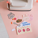 Children's craft project with paper cutouts of a birthday cake and bunnies on a pink surface.