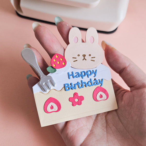 Hand holding a small decorative card with a rabbit on top of a cake, featuring 'Happy Birthday' text.
