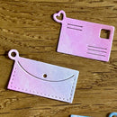 Two pink paper cutout shapes resembling a postcard and an envelope on a wooden surface.