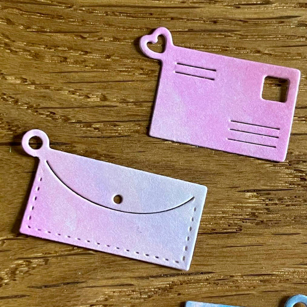 Two pink paper cutout shapes resembling a postcard and an envelope on a wooden surface.