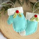 Two turquoise mitten ornaments with red berries and green leaves on a wooden surface.