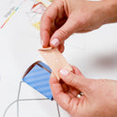 Person applying a small piece of paper with holes to a wooden stick, with a blurred background.