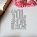 Metallic 'You are the world' cutout on a white surface with pink roses and an open book.