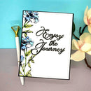 Notebook with floral design and 'Enjoy the Journey' text, accompanied by a pen and flowers on a blue and gray background.