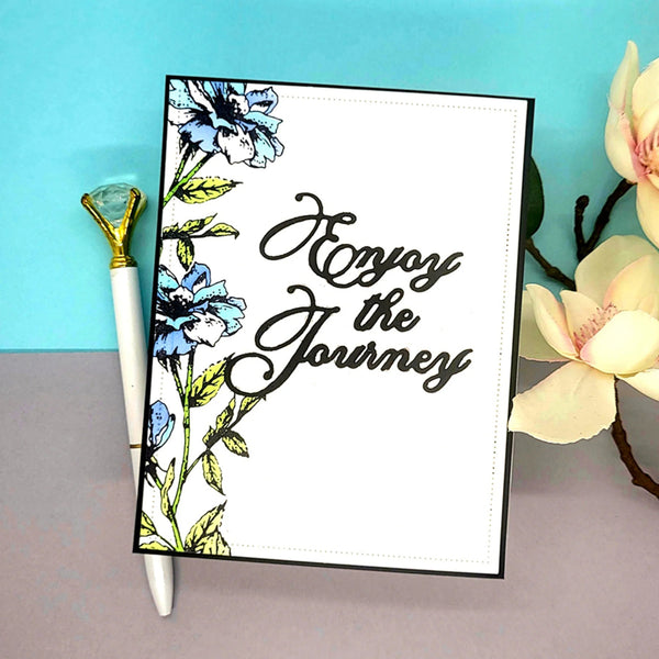 Notebook with floral design and 'Enjoy the Journey' text, accompanied by a pen and flowers on a blue and gray background.