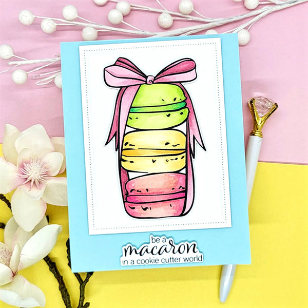 Card with colorful macaron illustration and 'be a macaron in a cookie cutter world' text on a pink and yellow background.