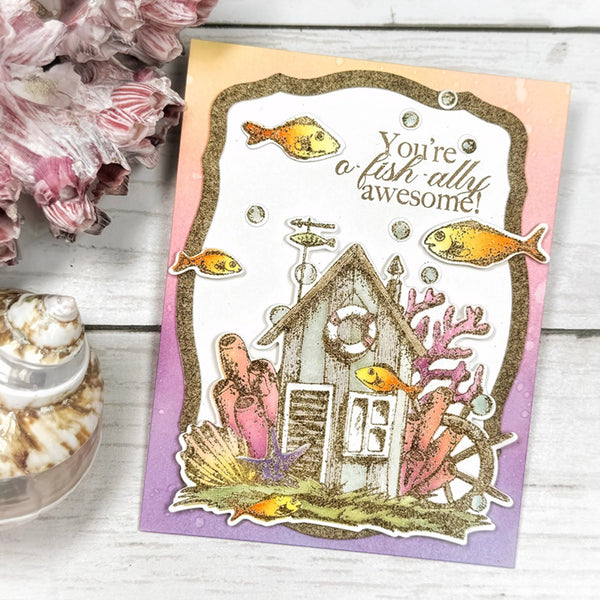 Underwater-themed card with fish and a house, placed on a wooden surface with flowers and shells.