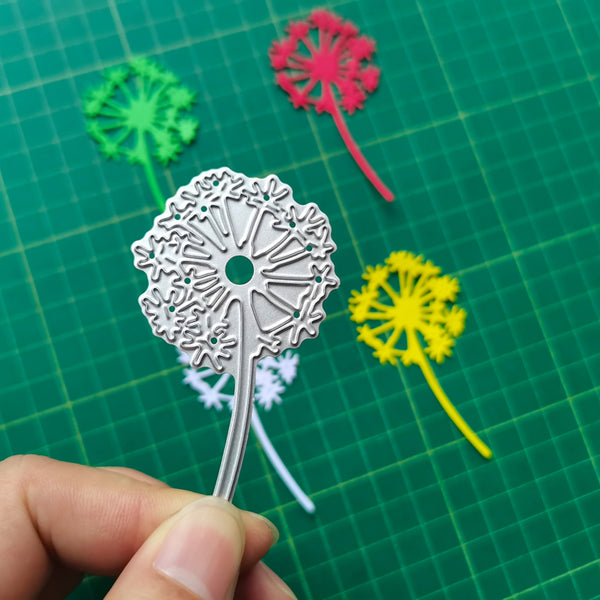 Metal dandelion flower cutout held by a hand on a green cutting mat with colorful paper dandelions.