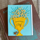 Card with yellow trophy design and 'World's Greatest Dad' text on a blue background, placed on a wooden surface.