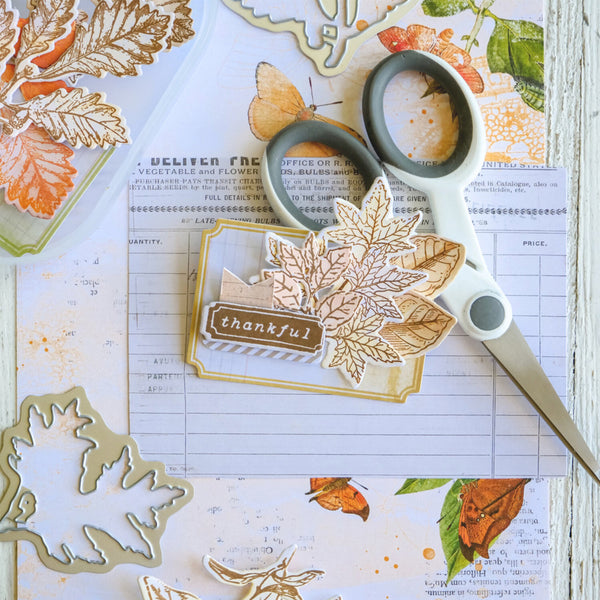 Card with autumn leaf design and 'thankful' text, surrounded by leaf cutters and scissors on a printed paper background.