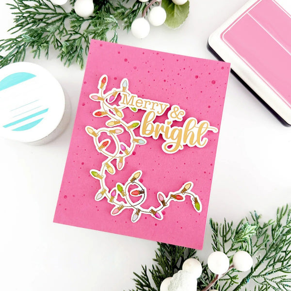 Pink card with 'Merry & Bright' text and Christmas lights design, surrounded by festive decorations on a white background.
