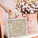 Decorative gift bag with intricate patterns, hand holding a small wrapped gift, and a basket of flowers in the background.