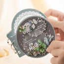 Decorative round object with floral and butterfly design held by a hand against a blurred background