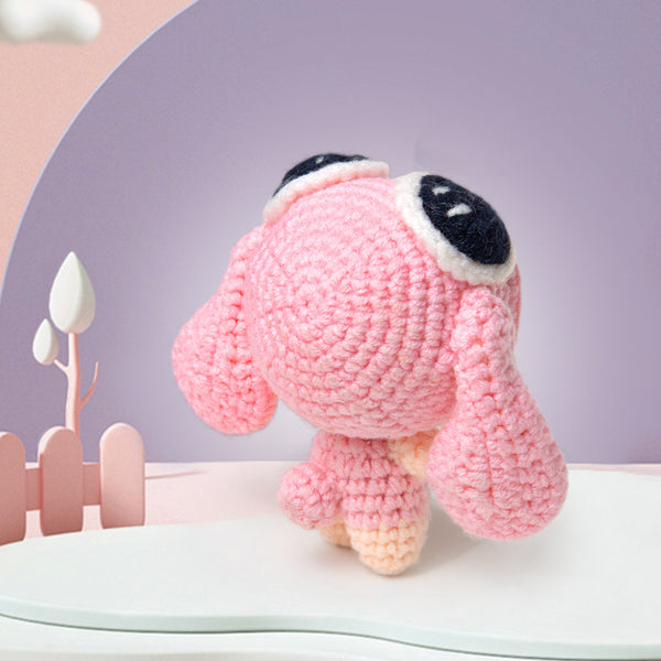 Pink crochet dog toy with black eyes on a white surface with a pink and gray background