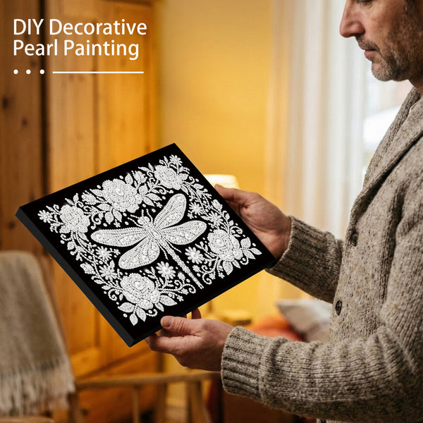 Man holding a black canvas with white decorative pearl painting of a dragonfly and flowers.
