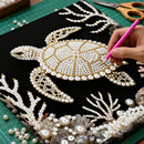 Handmade turtle design with pearls and gold accents on a black background, with a hand holding a pink tool.