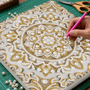 Hand applying gold and pearl embellishments to a decorative sheet on a green cutting mat.