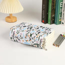 A fabric-covered notebook or planner with a colorful floral and bird pattern, shown on a white desk next to a pleated lamp, books, and colored pencils.