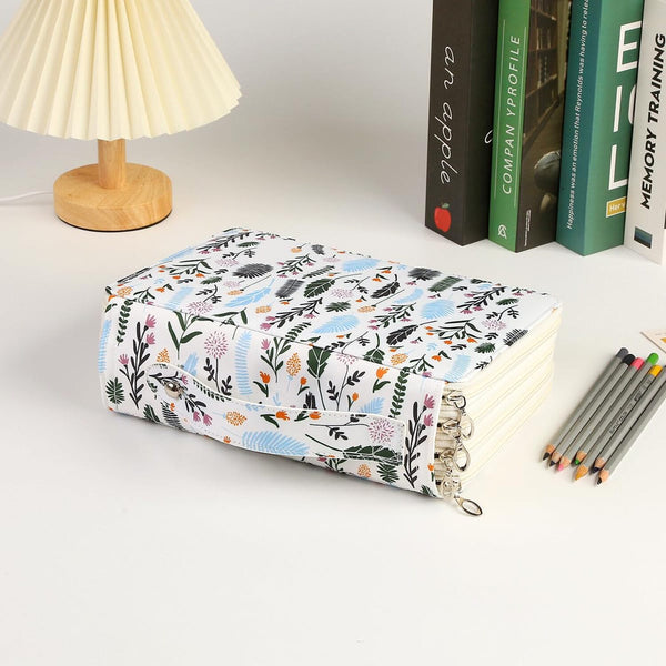 A fabric-covered notebook or planner with a colorful floral and bird pattern, shown on a white desk next to a pleated lamp, books, and colored pencils.