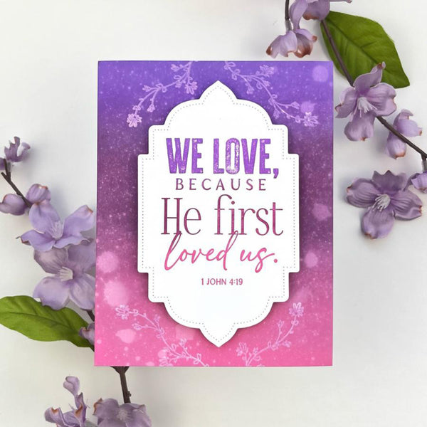 Card with 'We love, because He first loved us' text on a purple and pink floral background