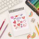 Notebook with heart and love drawings, colored pencils, and a keyboard on a white surface.