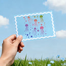 Hand holding a transparent sheet with colorful jellyfish illustrations against a blue sky and grass background