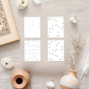 Four white cards with black patterns on a light wooden surface with decorative items.