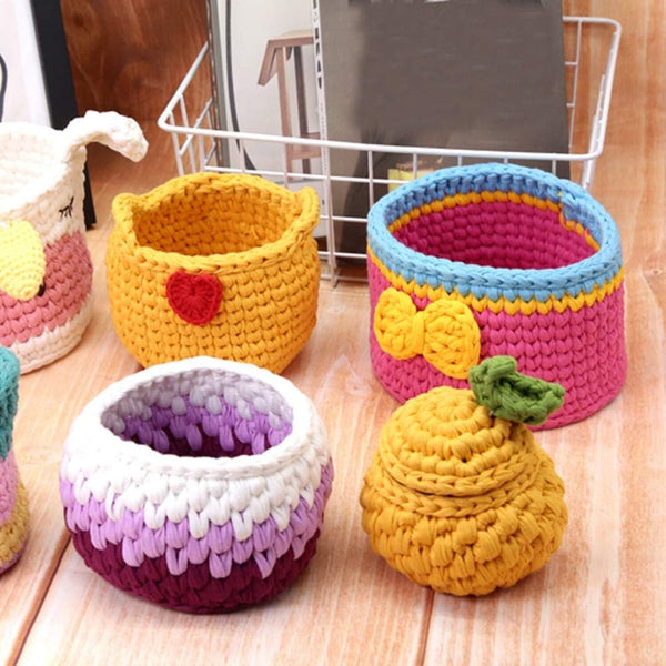 A set of handmade crocheted storage baskets and containers in various colorful patterns, including yellow with a red heart, pink and blue with a yellow bow, purple and white, and yellow with a green leaf, arranged on a wooden surface.