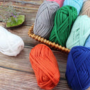 Multiple thick yarn balls in assorted colors arranged on a rustic wooden surface, including white, gray, green, mint, peach, orange, and blue. Some yarn balls are placed in a small woven basket, and a decorative green stone is visible in the background.
