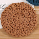 A round braided rope coaster in a warm tan-brown color, displayed on a light wood surface with a white paper card and a blue coaster partially visible in the background.