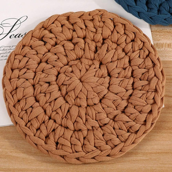 A round braided rope coaster in a warm tan-brown color, displayed on a light wood surface with a white paper card and a blue coaster partially visible in the background.